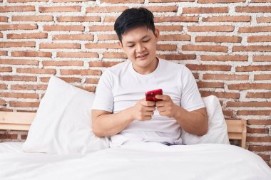 Young chinese man using smartphone sitting on bed at bedroom