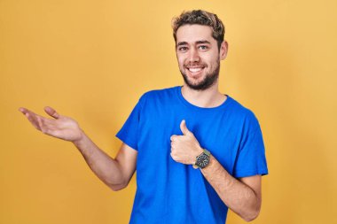Hispanic man with beard standing over yellow background showing palm hand and doing ok gesture with thumbs up, smiling happy and cheerful 