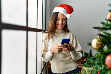 Middle age hispanic woman using smartphone standing by christmas tree at home