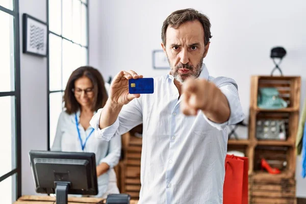 Hispanic man holding credit card at retail shop pointing with finger to the camera and to you, confident gesture looking serious