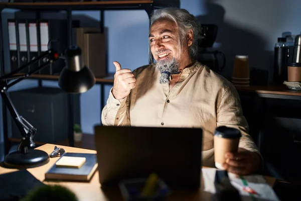 Middle age man with grey hair working at the office at night smiling with happy face looking and pointing to the side with thumb up.