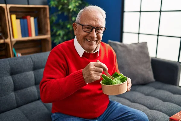 Senior man eating salad sitting on sofa at home