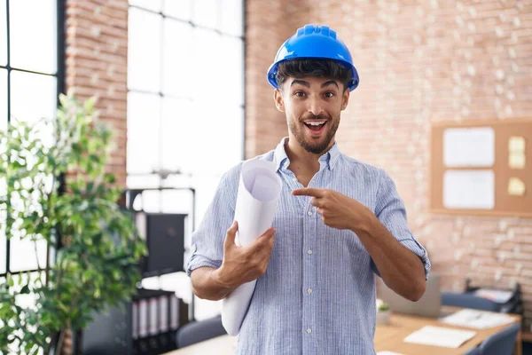 Arab man with beard wearing architect hardhat at construction office smiling happy pointing with hand and finger