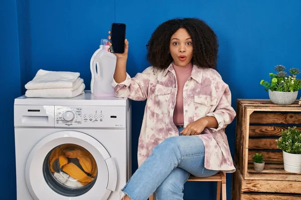 年轻的非洲裔美国女人用智能手机洗衣服时 吓得张大了嘴 脸上充满了惊奇和怀疑 — 图库照片