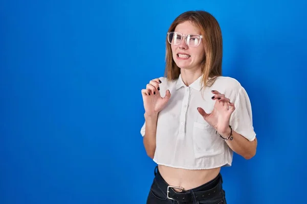 Beautiful woman standing over blue background disgusted expression, displeased and fearful doing disgust face because aversion reaction. with hands raised