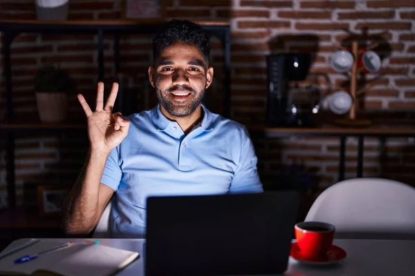 Hispanic man with beard using laptop at night showing and pointing up with fingers number three while smiling confident and happy.
