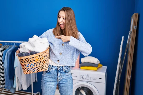 Young woman holding laundry basket smiling happy pointing with hand and finger