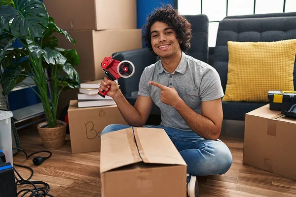 Hispanic man with curly hair moving to a new home closing cardboard box with tape smiling happy pointing with hand and finger
