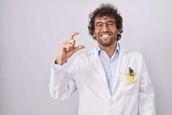 Hispanic young man wearing doctor uniform smiling and confident gesturing with hand doing small size sign with fingers looking and the camera. measure concept.