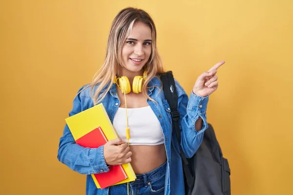 Young blonde woman wearing student backpack and holding books with a big smile on face, pointing with hand finger to the side looking at the camera.