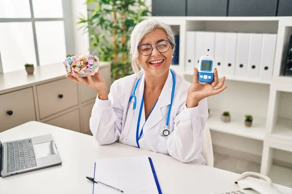 Middle age woman with grey hair wearing doctor uniform holding glucose monitor winking looking at the camera with sexy expression, cheerful and happy face.