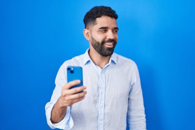 Hispanic man with beard using smartphone typing message looking away to side with smile on face, natural expression. laughing confident. 
