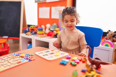Adorable hispanic girl playing with maths puzzle game sitting on table at kindergarten