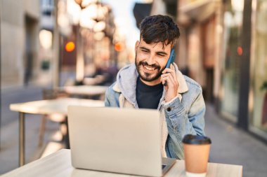 Young hispanic man using laptop talking on smartphone sitting on table at coffee shop terrace