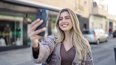 Genç sarışın kadın, kendinden emin gülümsüyor. Sokakta akıllı telefonun yanında özçekim yapıyor.