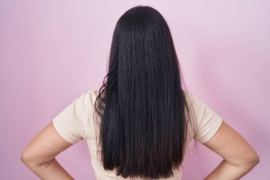 Young hispanic woman standing over pink background standing backwards looking away with arms on body 