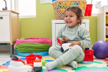 Adorable hispanic toddler playing with supermarket toy sitting on floor at kindergarten