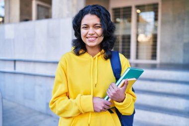 Genç Latin kız öğrenci gülümsüyor. Elinde kitaplarla.