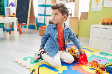 Anaokulunda yerde oturmuş traktör ve dino oyuncaklarıyla oynayan sevimli İspanyol bebek.