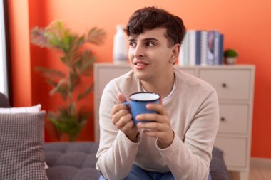 Non binary man drinking coffee sitting on sofa at home
