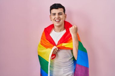 Non binary person holding rainbow lgbtq flag screaming proud, celebrating victory and success very excited with raised arms 