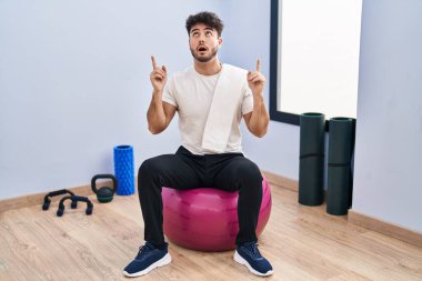 Hispanic man with beard sitting on pilate balls at yoga room amazed and surprised looking up and pointing with fingers and raised arms. 