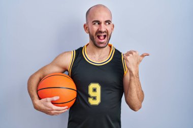 Young bald man with beard wearing basketball uniform holding ball smiling with happy face looking and pointing to the side with thumb up. 