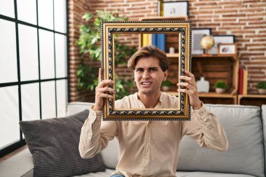 Young man holding empty frame clueless and confused expression. doubt concept. 
