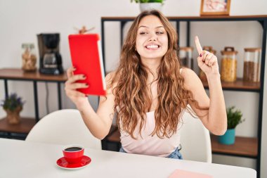 Young hispanic woman doing video call with tablet smiling with an idea or question pointing finger with happy face, number one 