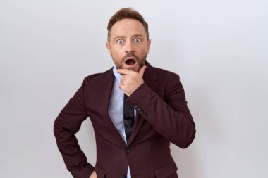 Middle age business man with beard wearing suit and tie looking fascinated with disbelief, surprise and amazed expression with hands on chin 