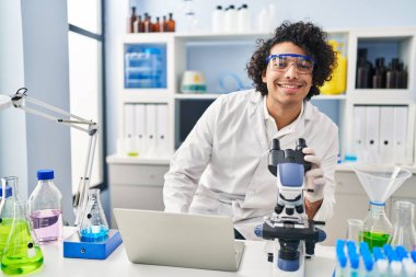 Laboratuvarda dizüstü bilgisayar ve mikroskop kullanan bilim adamı üniforması giyen genç bir İspanyol.