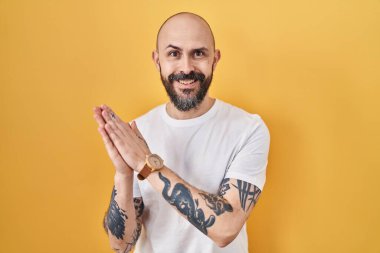 Young hispanic man with tattoos standing over yellow background clapping and applauding happy and joyful, smiling proud hands together 