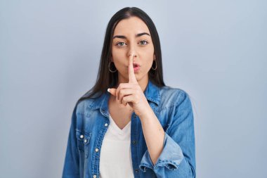 Mavi arka planda dikilen İspanyol kadın parmakları dudaklarında sessiz olmak istiyor. sessizlik ve gizli konsept. 