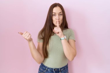 Beautiful brunette woman standing over pink background asking to be quiet with finger on lips pointing with hand to the side. silence and secret concept. 