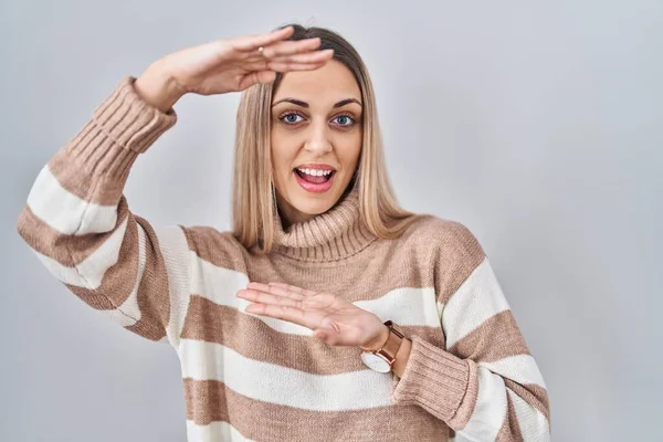Young blonde woman wearing turtleneck sweater over isolated background gesturing with hands showing big and large size sign, measure symbol. smiling looking at the camera. measuring concept.