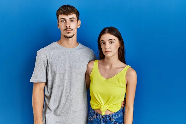 Young hispanic couple standing together over blue background relaxed with serious expression on face. simple and natural looking at the camera.