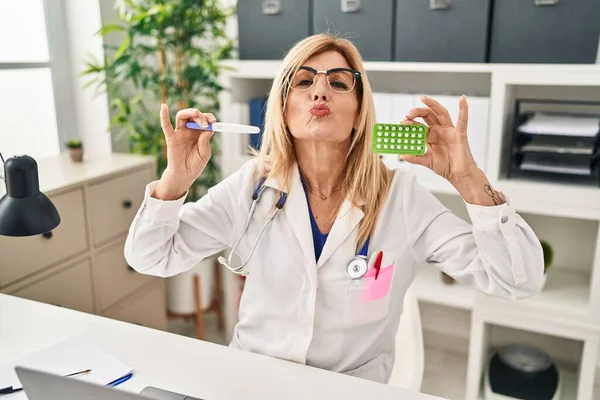 Middle age blonde doctor woman holding prescription pills and pregnancy test looking at the camera blowing a kiss being lovely and sexy. love expression.