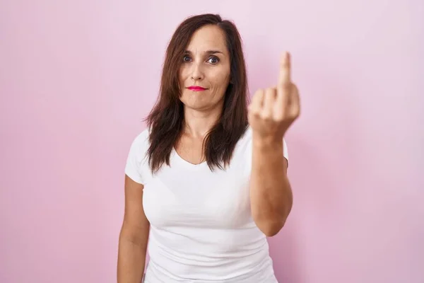 Middle age brunette woman standing over pink background showing middle finger, impolite and rude fuck off expression