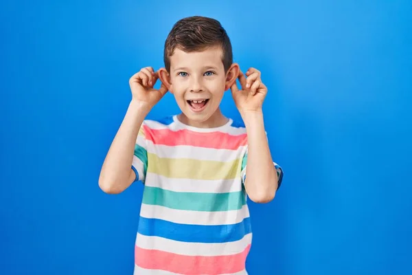 年轻的高加索小孩站在蓝色的背景上 微笑着用手指拉耳朵 滑稽的手势 试镜问题 — 图库照片