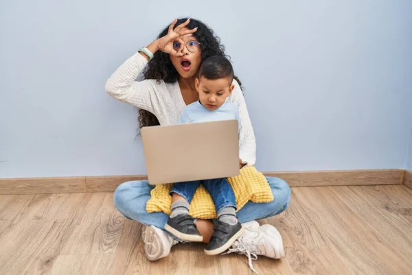 Young hispanic mother and kid using computer laptop sitting on the floor doing ok gesture shocked with surprised face, eye looking through fingers. unbelieving expression.