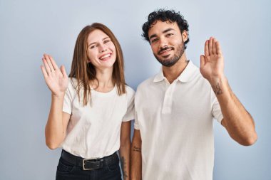 Young couple wearing casual clothes standing together waiving saying hello happy and smiling, friendly welcome gesture 
