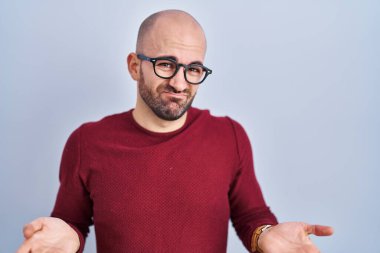 Young bald man with beard standing over white background wearing glasses clueless and confused with open arms, no idea concept. 