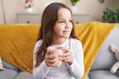 Adorable hispanic girl drinking cup of milk sitting on sofa at home