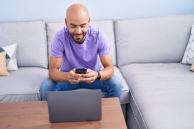Young bald man using smartphone and laptop at home