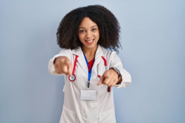 Genç Afro-Amerikan kadın doktor üniforması ve steteskop takmış parmaklarıyla sizi ve kamerayı işaret ediyor. 