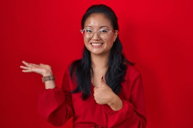 Asian young woman standing over red background showing palm hand and doing ok gesture with thumbs up, smiling happy and cheerful 