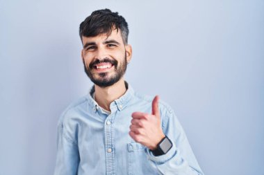 Young hispanic man with beard standing over blue background doing happy thumbs up gesture with hand. approving expression looking at the camera showing success. 