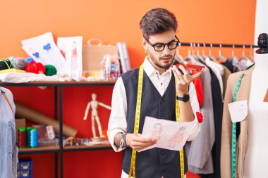 Young hispanic man tailor talking on smartphone looking clothing design at atelier
