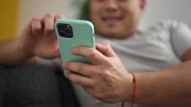 Young chinese man using smartphone sitting on sofa at home