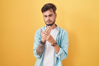 Young hispanic man with tattoos standing over yellow background suffering pain on hands and fingers, arthritis inflammation 